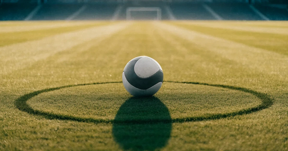 Uma bola de futebol no ponto central de um relvado, à luz da manhã